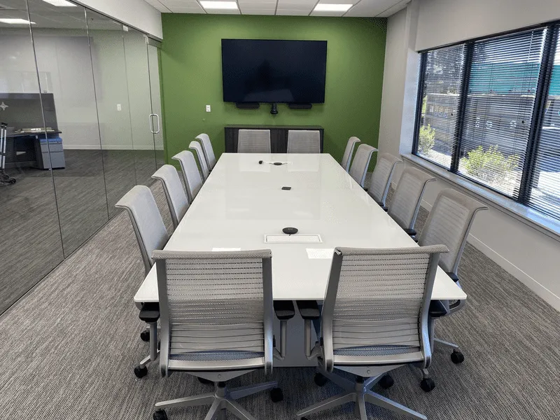 A modern conference room featuring a long table surrounded by ergonomic task chairs, with a TV mounted on the back wall.