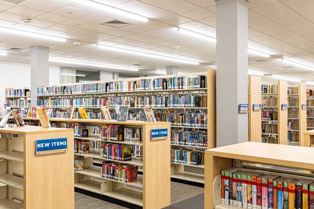 Library bookcases filled with books, featuring signage indicating the contents of each shelving unit.